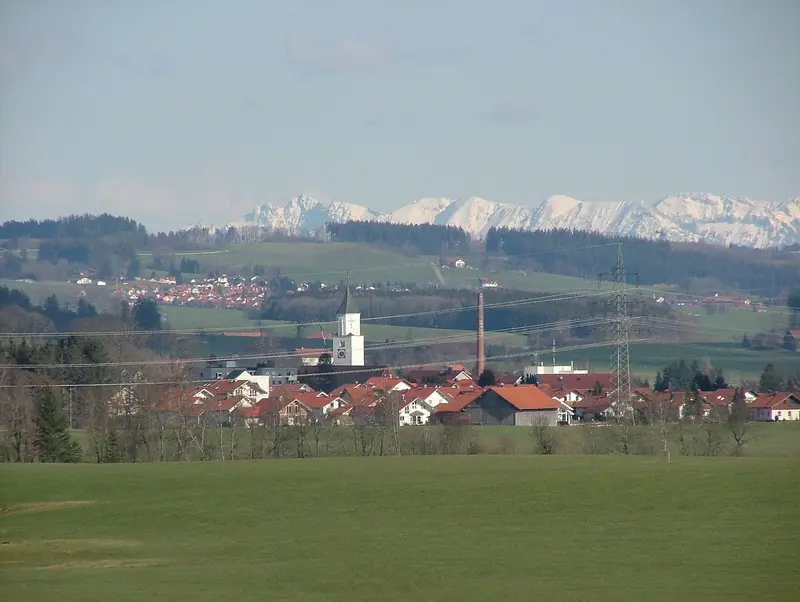 Blick über Felder auf Dietmannsried mit Kirchturm, dahinter die schneebedeckten Allgäuer Alpen