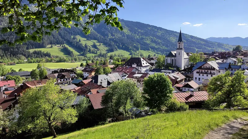 Blick über die Dächer von Bad Hindelang mit der Pfarrkirche St. Johannes Baptist und bewaldeten Bergen im Ostrachtal