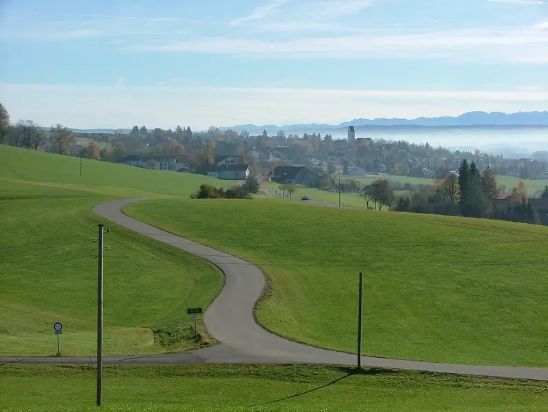 Blick über grüne Wiesen und eine geschwungene Straße auf Altusried mit Kirchturm, im Hintergrund die Allgäuer Alpen
