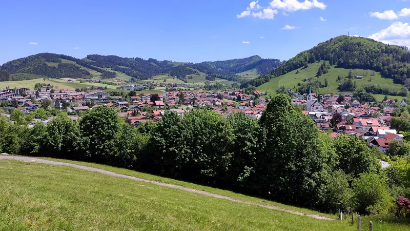Blick von Westen auf Oberstaufen mit der Pfarrkirche St. Peter und Paul, umgeben von grünen Hügeln und Bergen